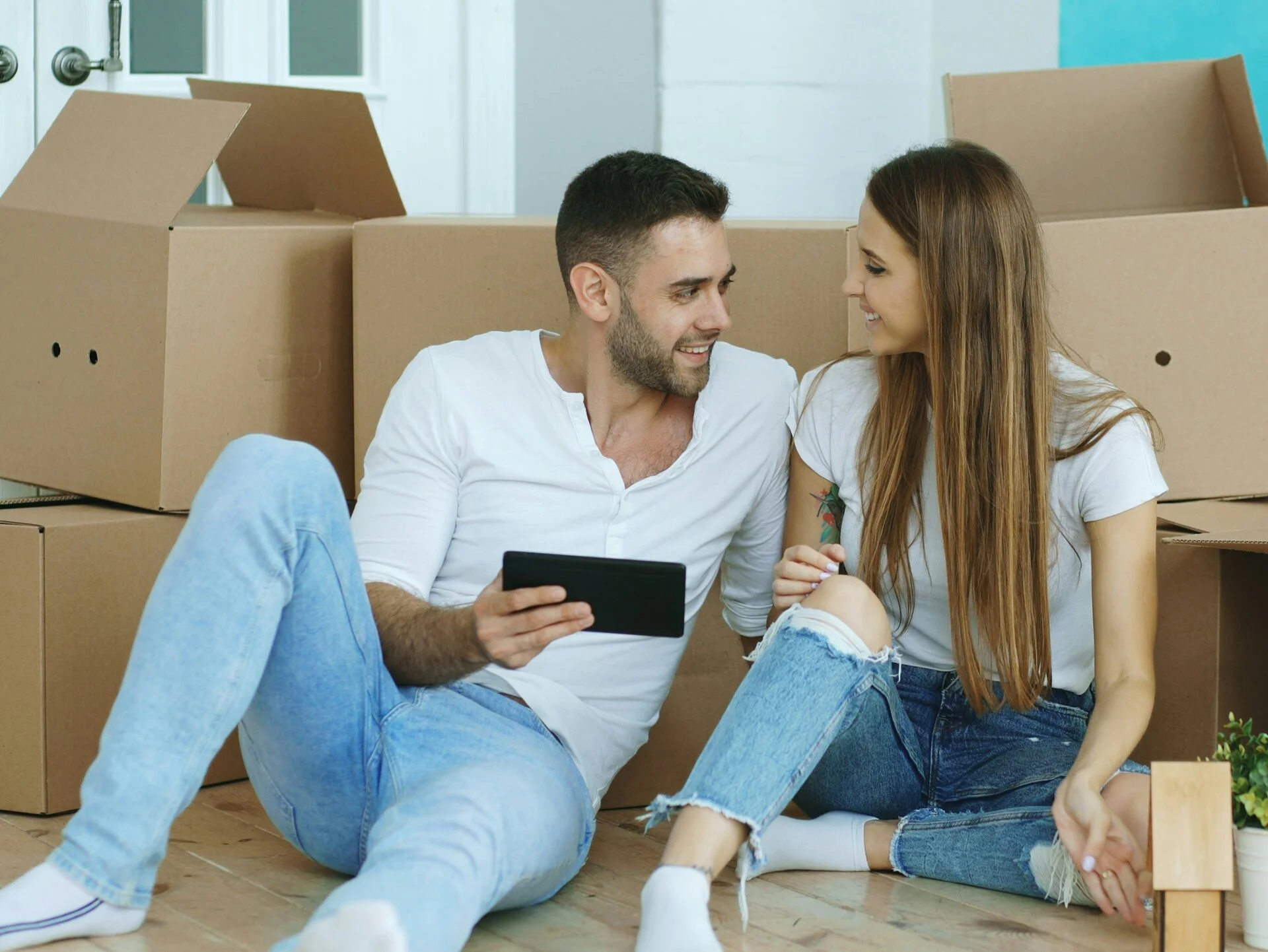 Couple sitting on floor surrounded by moving boxes, planning their new home setup on a tablet